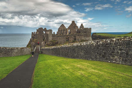 Tourist walking to main entrance of Dunluce castle. Northern Ireland. Close-up path to the Medieval ruins. Grass covered lawns before the now-ruined historical building. Picturesque Irish shoreline.の写真素材