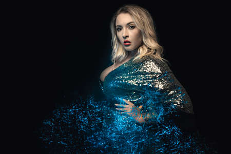 Close-up woman blonde looking toward the camera. Mixed lights effects in blue tints. Black background. Posing young woman in decollete evening dress with sequins. Plus size model.の写真素材