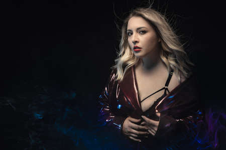 Close-up blonde in black bra and lacquer raincoat. Mix lights effect. Portrait of young woman in sexual outfit on the black background. Blue fog surrounding. Sensual fashion shot.の写真素材