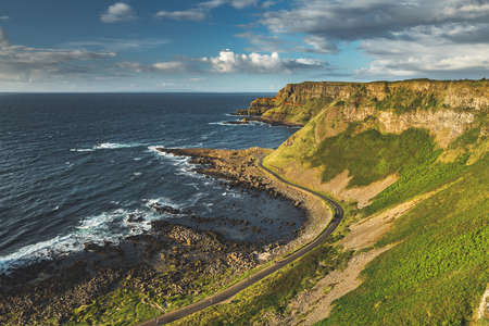 Coast Scenery Nothern Ireland Panoramic Photo. Beautiful View on Waves Sea Bay, Asphalt Road, Green Grass Climb and Sheer Cliff. Warm Shiny Day, Blue Sky with Clouds. National Country Tourist Placeの写真素材