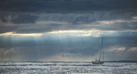 Elegant Touristic Yacht in Ocean Northern Ireland. Stormy Clouds, Sun Rays and Island on Horizon. Outdoor Adventure Activity. Panoramic Photo with Lagoon, Boat and Island on Backgroundの写真素材