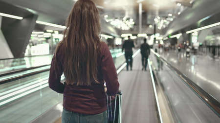 Woman with Luggage Stand on Speedwalk at Airport. Back View of Caucasian Girl with Trolley. Female Tourist on Moving Travelator. People Walking by. Modern Depature Terminal.の写真素材