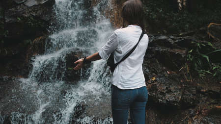 Woman Tourist Reaching out Hands to Splashing Waterfall. Caucasian Young Girl Wearing Jeans and Shirt Hike at Beautiful Canyon. Traveler Enjoy Cool Water Flood. Tropical Vacation.の写真素材