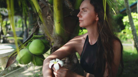 Young Carefree Woman Sitting under Palm Tree Leaf. Beautiful Tanned Girl Sniff Tropical Flower. Tourist Enjoy Exotic Vacation. Koh Phangan Island Resort, Thailand.の写真素材