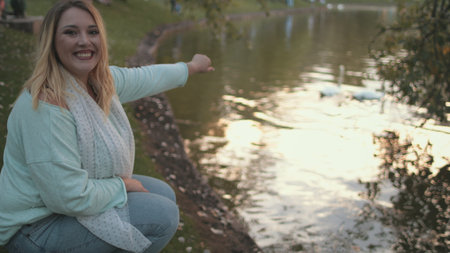 Pretty Woman Plus Size Laugh Relax At Lake Surface. Happy Caucasian Girl Clap Hands Look at Swan Couple City Park River Background. Young Blonde Lady Smile Wear Casual Clothesの写真素材