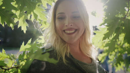Nice Woman Plus Size Smile Stand By Park Tree. Blonde Caucasian Girl Enjoy Greenish Leaves City Park Nature Background. Joyful Overweight Female Adult Sunbeam Summertime Happy Momentsの写真素材