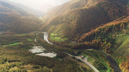 grey asphalt road along blue winding mountain river surrounded by brown dense forests lit by bright sun aerial view. Carpathian mountains, Ukraine beauty nature. Travel, summer holidays.の写真素材