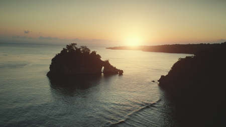 Aerial Drone Flight from Rock Mountain Arch in Sunset Ocean Water. Beautiful Wild Beach Bay on Tropical Nusa Penida Island, Indonesia. Nature Landscapeの写真素材