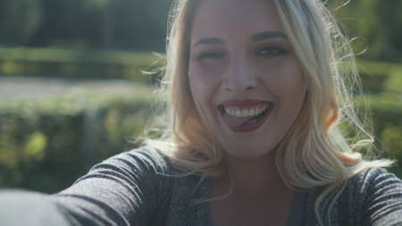 Pretty Blonde Girl Give Wink Take Selfie Closeup. Attractive Caucasian Woman Plus Size Laugh Rotation Around City Park Background. Happy Female Adult Smile Tongue Happiness Conceptの写真素材