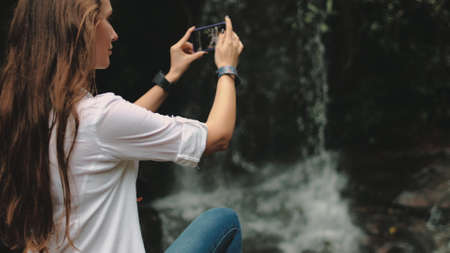 Young Woman Traveler near Waterfall at Rainforest. Caucasian Girl make Video of Mountain Cascade Stream. Female Hiker Holding Smartphone. Exotic Thailand Vacation.の写真素材