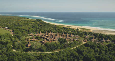 Aerial traditional village with ornated roofs houses at sea sand coast. Indonesia tourist attraction at green scape on ocean bay. Cinematic settlement landmark of Sumba Island, Asiaの写真素材