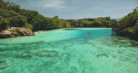 Closeup people swim at turquoise crystal clean water of Weekuri lake. Tourists landmark in green tropic forest with high bridge. Magical summer vacation in tropical paradise of Sumba Island, Indonesiaの写真素材