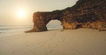 Sand ocean beach at sun light with cliff shore aerial view. Giant hole at rock wall with green grass top. Sumba island landmark Batu Bolong, Indonesia tourist attraction at nobody nature sceneの写真素材
