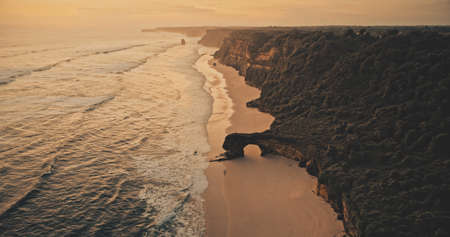 Sunset ocean seascape with waves at rock wall on sand beach aerial view. Nobody nature scenery at summer evening with amazing seascape at cliff sea shore. Soft light Indonesia sun set scapeの写真素材