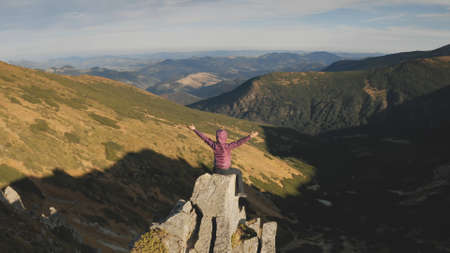 Woman rising up hands aerial. Tourist girl sit on mountain rock top. Nature landscape at autumn day. Mount hills with grass. Mountaineering lifestyle. Carpathians ridges, Ukraine, Europe. Drone shotの写真素材