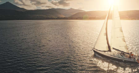 Sun reflection at sailboat on ocean bay aerial. Summer sea cruise on ship at green mountains island of Arran on yachts with passengers. Scotland sea coast water with vessel. Cinematic drone shotの写真素材