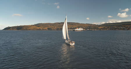 Slow motion of yacht reflection at sea gulf aerial. Alone sail boat reflect at ocean bay. Racing sailboat at Brodick Harbor, Scotland, Europe. Luxury vessel at summer cruise. Cinematic soft light shotの写真素材