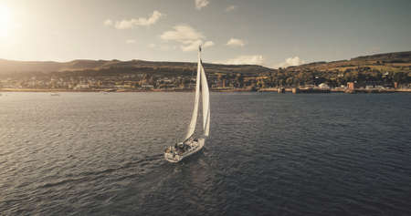 Aerial view of sun light on sail boat at ocean bay reflection. Highland sea coast with forest and mounts at bright sunlight. Nobody nature seascape at Brodick harbor with luxury yachts cinematic shotの写真素材