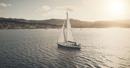 Sun seascape with white sailboat aerial. Mesmerezing landscape with sail boat at summer. Sunlight over sail boat at ocean bay. Sea shore of Arran Island with vessel. Serene water scenery drone shotの写真素材