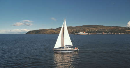 Sailboats cruise at open sea. Brodick port, Arran Island, Scotland. Nature seascape at summer day. Yachting at cargo terminal on ocean harbor aerial. Regatta on luxury yacht. Cinematic drone shotの写真素材
