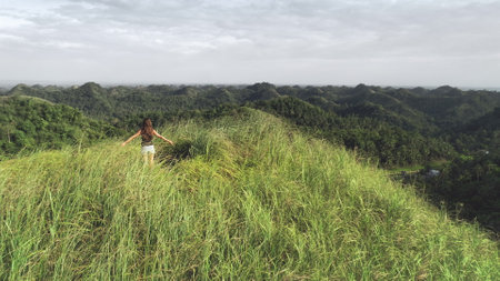 Woman rising up hands aerial view: green grass mount top in tropical Philippines. Traveler girl on hill grassy peak fill freedom of wild asian nature. Epic landcape of exotic mountains in drone shotの写真素材