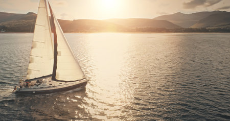Sun reflection at sailboat on ocean bay aerial. Summer sea cruise on ship at green mountains island of Arran on yachts with passengers. Scotland sea coast water with vessel. Cinematic drone shotの写真素材