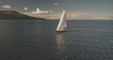 Sail boat at mountain ocean shore of Scotland Island of Arran aerial. Yacht sails with beautiful fluffy clouds sky at open sea. Sailboat cruise at summer sun shine day. Cinematic soft light drone shotの写真素材