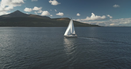 Slow motion of yacht, wind blows to sail at ocean bay aerial. Mountain island of Arran, Scotland, Europe. Nobody nature seascape at Brodick Gulf. Luxury cruise on sailboat. Cinematic tourism lifestyleの写真素材