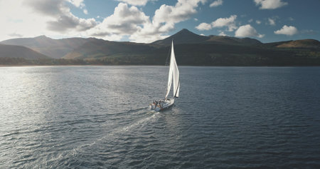 Slow motion of yacht, wind blows to sail at ocean bay aerial. Mountain island of Arran, Scotland, Europe. Nobody nature seascape at Brodick Gulf. Luxury cruise on sailboat. Cinematic tourism lifestyleの写真素材