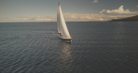 Sail boat at mountain ocean shore of Scotland Island of Arran aerial. Yacht sails with beautiful fluffy clouds sky at open sea. Sailboat cruise at summer sun shine day. Cinematic soft light drone shotの写真素材