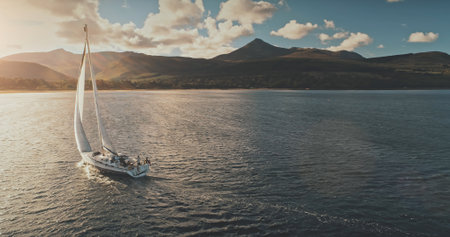 Sailing yacht in wind waves at Arran Island sea bay in Scotland. Amazing seascape at Brodick port city. Lonely sail boar race at sun shine with fluffy clouds. Serene water scape and transportationの写真素材