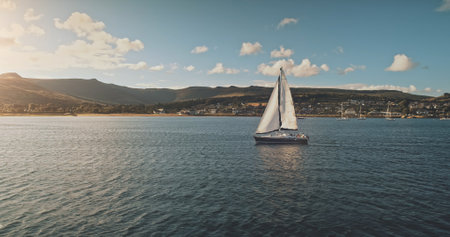 Sailing at racing yacht in open sea, sun shine aerial view. Majestic seascape at Arran Island coast with blue sky background. Hilly isle of Scotland with Brodick pier. Luxury lifestyle cinematic sceneの写真素材