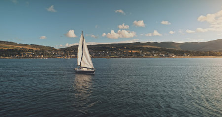 Sailing at racing yacht in open sea, sun shine aerial view. Majestic seascape at Arran Island coast with blue sky background. Hilly isle of Scotland with Brodick pier. Luxury lifestyle cinematic sceneの写真素材