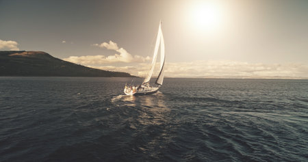 Aerial panorama of yacht race in open sea with sun reflections. Serene seascape with sailing boat. Luxury yachting at ocean bay water. Cinematic water transport scenery at summer cruise conceptの写真素材
