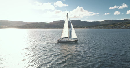 Slow motion of sun ocean gulf with yacht sailing aerial. Sunlight reflection at sea bay. Sailboat at Arran coast, Scotland, Europe. Luxury vessel at summer cruise. Cinematic soft light shotの写真素材