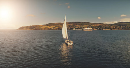 Slow motion of sun ocean gulf with yacht sailing aerial. Sunlight reflection at sea bay. Sailboat at Arran coast, Scotland, Europe. Luxury vessel at summer cruise. Cinematic soft light shotの写真素材