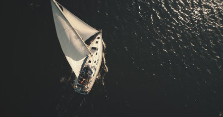 Top down of yacht under white sail at dark open sea aerial. Ocean bay black water and lonely sailboat. Summer cruise on boat. Luxury yachting scenery at serene seascape in dramatic drone shotの写真素材