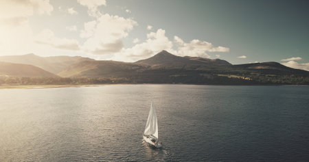 Sun shine over ocean bay with sailing yacht reflection aerial. Amazing sunlight at summer luxury cruise on sail boat at open sea. Mountain island with green forest at coast. Fluffy clouds over mountの写真素材