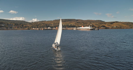 Port town with cargo terminal and yachts on ocean at harbor of Brodick. Sail boat at sea gulf. Cruise at passenger ship race at bay with highlang island shore. Sun reflect at sailboat. Epic seascapeの写真素材