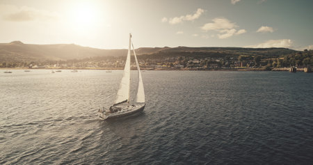 Aerial view of sun light on sail boat at ocean bay reflection. Highland sea coast with forest and mounts at bright sunlight. Nobody nature seascape at Brodick harbor with luxury yachts cinematic shotの写真素材