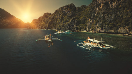 Seascape with passenger boats at sunset in closeup aerial view. Cliff coast with green trees at sunny summer day. Philippines vacation with tourists swim at ocean water. Sun set cinematic drone shotの写真素材