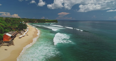 Stunning aerial drone highlights Dreamland Beach in Bali, Indonesia, with turquoise waves gently crashing on the sandy shore, vibrant beach umbrellas, and a red beach hutの写真素材