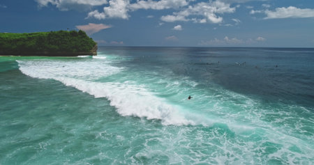 Turquoise ocean waves rolling towards Dreamland Beach in Bali, Indonesia, with surfers riding the waves and a rocky cliff covered in lush green vegetation in the background under a cloudy skyの写真素材