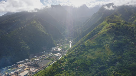 Aerial view of a lush green valley in Tahiti, French Polynesia, with sun rays penetrating through clouds, illuminating a river flowing amidst the mountains and industrial buildingsの写真素材
