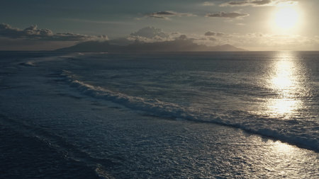 Golden sunset light reflects on the surface of a calm ocean as gentle waves break near a tropical island, creating a serene and idyllic sceneの写真素材