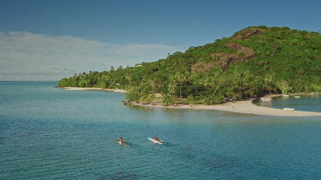 Two polynesian men paddling outrigger canoes near tropical island shore with lush vegetation and white sand beach in French Polynesia, aerial drone viewの写真素材