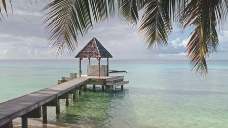French Polynesia, Rangiroa island: Palm tree leaves framing wooden pier and gazebo over turquoise lagoon coastal area, clear waters reflect the sky, creating a peaceful atmosphere ideal for relaxationの写真素材