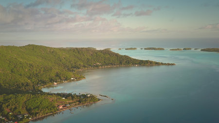 Aerial view of tropical island coastline showing lush green vegetation meeting calm turquoise ocean water at sunrise with small islands visible in the distanceの写真素材