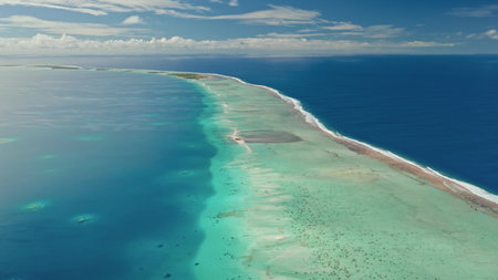 Crystal clear turquoise waters stretch out around a narrow sandy islet under a sunny sky. The coastline features stunning coral reefs that teem with aquatic life.の写真素材