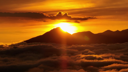 Mount Batur volcano silhouetted against a vibrant golden sunrise, with a sea of rolling clouds filling the valley below, creating a serene and breathtaking aerial landscape panoramaの写真素材